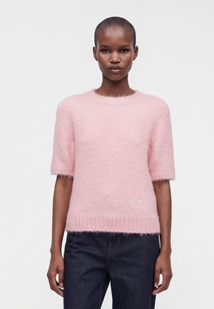 Femme aux cheveux courts portant un pull à manches courtes rose pâle et un jean bleu foncé sur un fond clair uni.