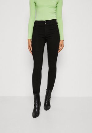 Pantalon noir slim fit avec fermeture double bouton, porté avec des bottines noires à bout pointu et un haut à manches longues vert clair.