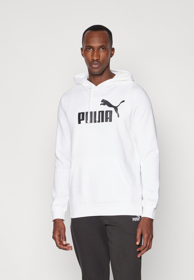 Man wearing white Puma hoodie with black logo and black Puma sweatpants, standing against a plain light background.
