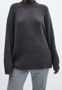 Een persoon die een donkergrijze fijngebreide trui met lange mouwen en lichtgrijze broek draagt, staand tegen een effen lichte achtergrond.