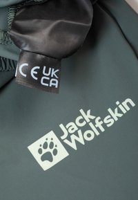 Donkergroene stof met een glanzend zwart label. Bevat het "Jack Wolfskin"-logo in een lichte kleur en de wettelijke aanduidingen "CE" en "UKCA."