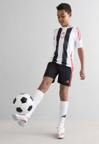 Gutt i svart og hvitt fotballdrakt som balanserer en svart-hvit fotball på venstre fot mot en enkel hvit bakgrunn.