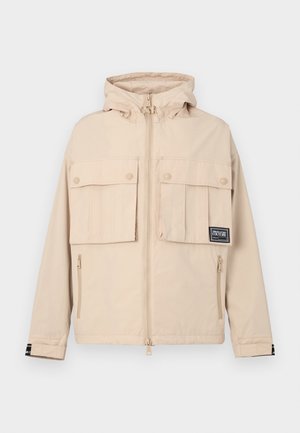 Beige hooded jacket with two front flap pockets and a side zipper pocket, featuring a black logo patch and a smooth, water-resistant fabric.
