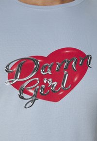 T-shirt en coton bleu clair avec un grand motif de cœur rouge et le texte « Damn Girl » en écriture argentée. Texture lisse, col rond.