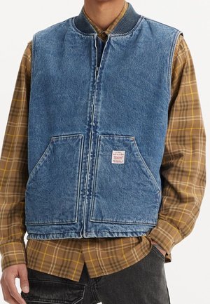 Homme portant une veste en jean bleue avec des poches avant sur une chemise à carreaux marron et un pantalon noir, debout avec une main dans la poche.