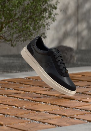 Zapatilla negra de corte bajo con suela blanca flotando sobre suelo de baldosas de madera, con follaje verde y fondo de pared gris.