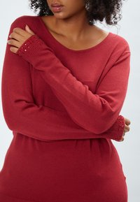 Maglione rosso in maglia con una vestibilità oversize. Presenta polsini a coste decorati e un ampio scollo. Tessuto morbido, design leggero.