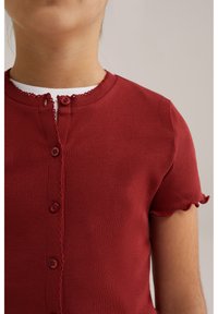 Camicetta rossa a righe con maniche corte, colletto rotondo, bordi decorativi a scallop e quattro bottoni abbinati sul davanti.