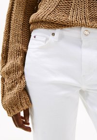 Vaqueros de denim blanco con un diseño ajustado y un pequeño detalle del logo en rojo, blanco y azul, combinados con un suéter de punto marrón texturizado.