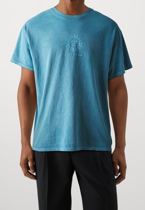 Otoman morska zielona bawełniana koszulka z krótkim rękawem, z haftowanym wzorem słońca i napisem "ŻYJ Z SŁOŃCEM", okrągły dekolt i luźny krój.