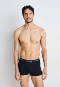 Zwarte katoenen boxershorts met een Calvin Klein logo op de tailleband. Het model is blote bovenlichaam en staat voor een neutrale achtergrond met natuurlijke verlichting.