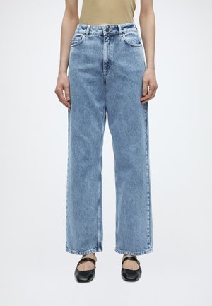 Személy világoskék, magas derekú, egyenes szárú farmernadrágot visel első zsebekkel, bézs felsővel és fekete zárt orrú, pántos cipővel párosítva.