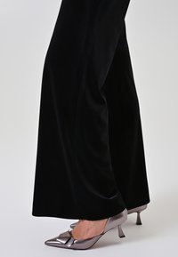 Scarpe con tacco alto color grigio metallizzato con dettaglio a fiocco, contrastate da pantaloni neri in velluto fluente che mostrano una texture liscia e una vestibilità sartoriale.