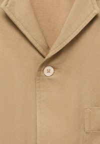 Camicia beige con tessuto texturizzato, caratterizzata da un colletto a punta, chiusura con un bottone singolo e dettaglio della tasca a sinistra.