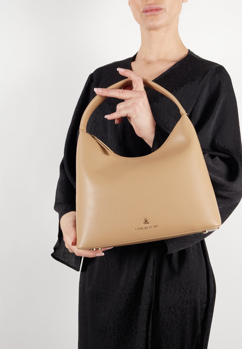 Torebka skórzana w kolorze tan, o zaokrąglonym kształcie i górnym uchwycie. Posiada złoty akcent z logo oraz gładką teksturę. Noszona z czarnym zestawem.