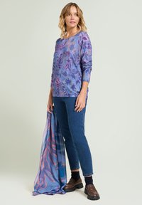 Maglione leggero viola a fantasia floreale con maniche lunghe, abbinato a jeans blu scuro e una sciarpa coordinata, tenuta in mano dalla modella.