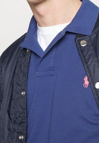 Marinblå polo med strukturerad väv och en rosa broderad logotyp, lager på lager under en mörk puffjacka med tryckknappar.