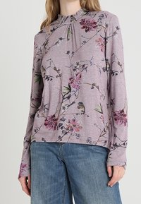 Långärmad lila skjorta med blommönster, med en insydd halsringning och mjukt tyg. Knappt med ljusblå denimjeans.