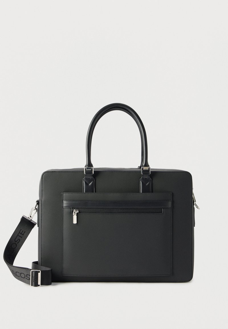 Mallette rectangulaire noire avec poignées supérieures, poche zippée à l'avant et bandoulière détachable avec le logo Lacoste.
