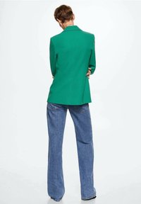 Vihreä räätälöity bleiseri, joka on valmistettu sileästä kankaasta ja yhdistetty vaaleansinisiin leveälahkeisiin farkkuihin. Bleiserissä on yksittäinen halkio takana.