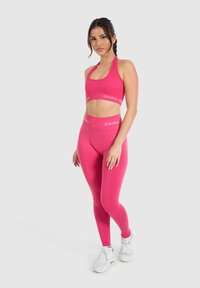 Top corto deportivo rosa y leggings con una banda de logo a juego. La modelo lleva zapatillas blancas, mostrando un diseño ajustado y elástico.