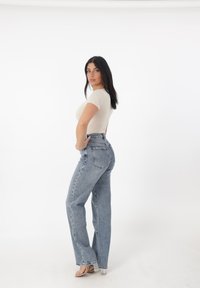 Top ajustado beige y jeans de tiro alto de color azul claro con pierna recta. La modelo posa de lado sobre un fondo blanco liso.