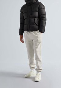 Doudoune noire avec un col montant, des poches zippées et des poignets côtelés, associée à un pantalon de jogging gris clair et des baskets blanches.