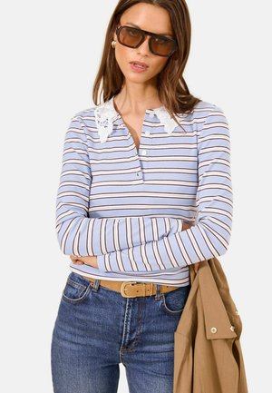 Naine, kes kannab pruunikate läätsedega päikeseprille, sinise ja valge triibulise pika varrukaga särki valge pitskraega, siniseid teksaseid ning hoiab käes beežikat jopet.