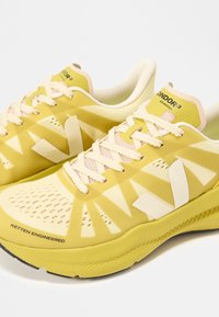 Ζευγάρι κίτρινων και εκρού παπουτσιών για τρέξιμο με λευκά κορδόνια, γεωμετρικά σχέδια και εμφανές το κείμενο "ONDR 3 ADVANCED" και "KETTEN ENGINEERED".