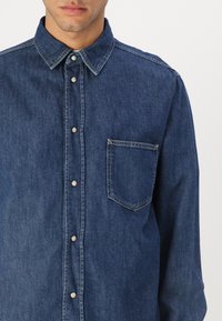 Camisa de mezclilla azul con cuello puntiagudo, botones a presión, mangas largas y un bolsillo en el pecho. Presenta una textura ligeramente deslavada.