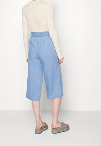 Mujer vestida con pantalones cortos de mezclilla de tiro alto y pierna ancha en azul claro, blusa de manga larga de canalé en beige y sandalias de plataforma beige, de pie contra un fondo blanco.