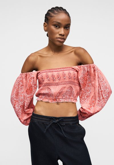 Femme aux cheveux tressés portant un haut court rose à épaules dénudées avec un imprimé cachemire et des manches volumineuses, associée à un pantalon foncé à cordon de serrage.