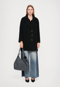 Manteau en laine noire mélangée avec de grands boutons et deux poches, associé à un jean en denim à jambe large. Un sac slouch en cuir gris vient compléter la tenue.