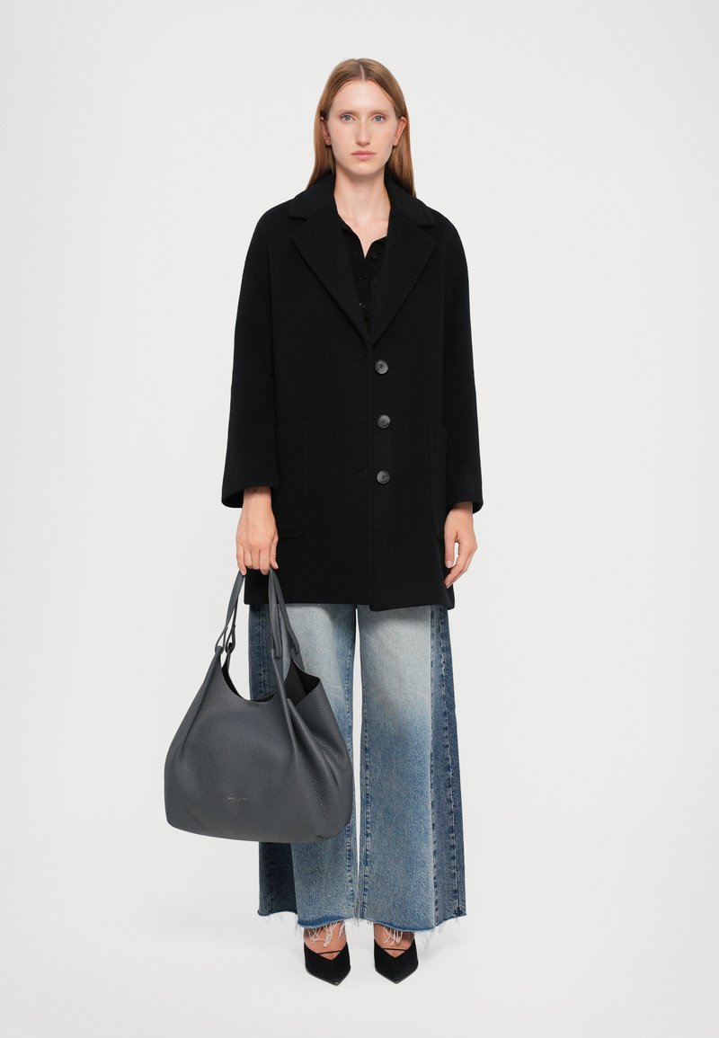 Manteau en laine noire mélangée avec de grands boutons et deux poches, associé à un jean en denim à jambe large. Un sac slouch en cuir gris vient compléter la tenue.