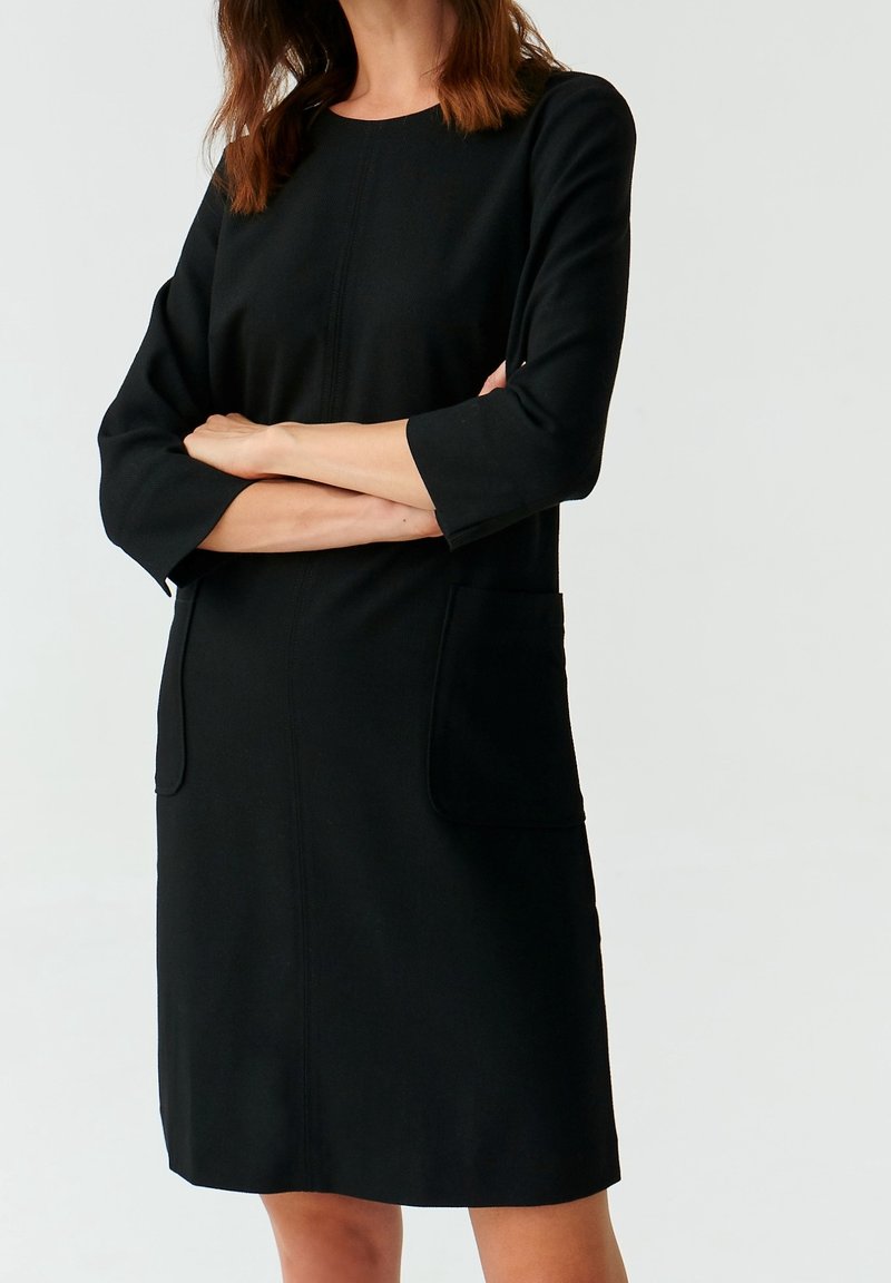 Femme portant une robe noire mi-longue aux genoux avec des manches trois-quarts et des poches avant, bras croisés devant un fond clair uni.