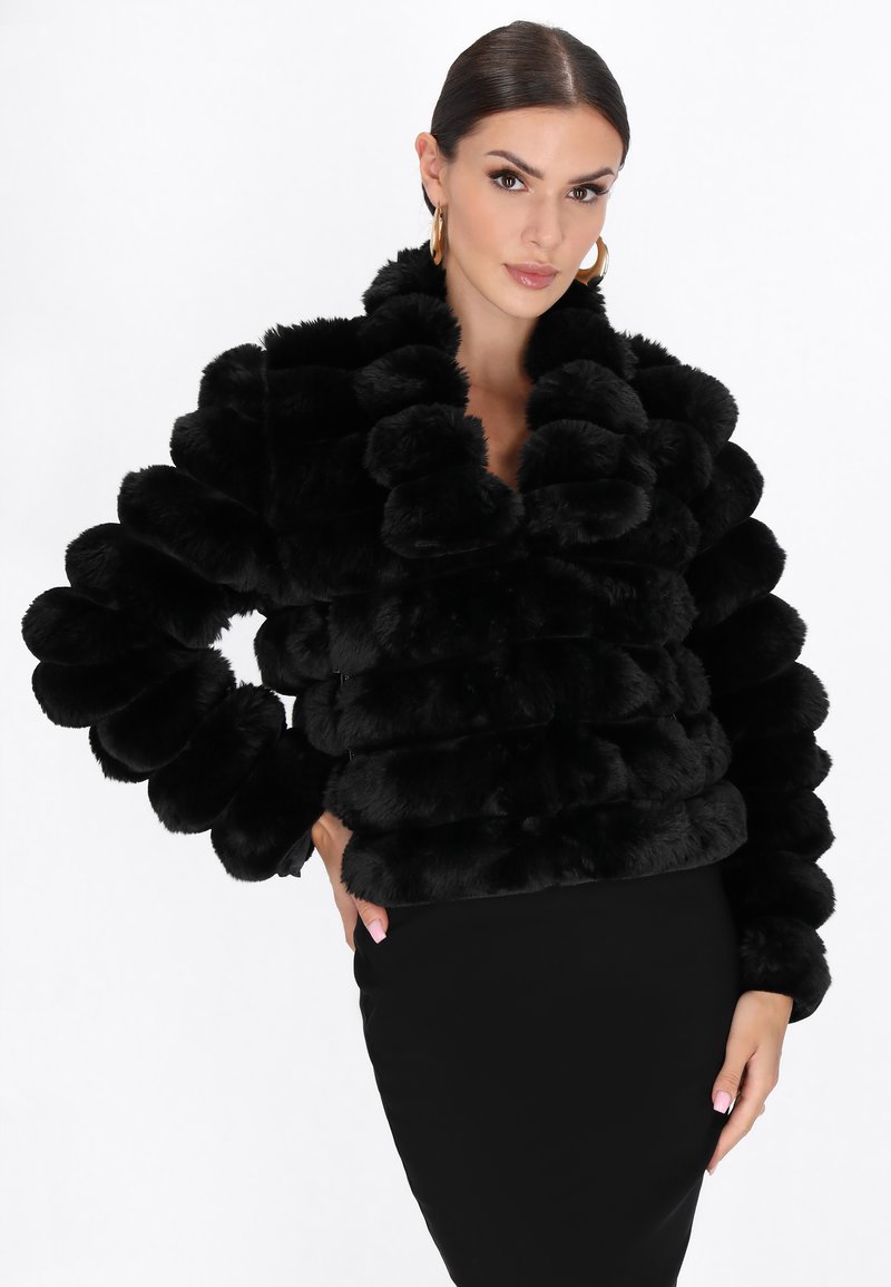 Frau mit nach hinten gegeltem Haar, die große goldene Creolen, eine schwarze strukturierte Fake-Fur-Jacke und einen taillierten schwarzen Rock trägt.