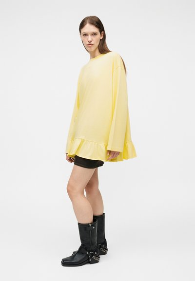Νεαρή γυναίκα με μεγάλο κίτρινο τοπ μακρυμάνικο με βολάν στο τελείωμα, μαύρα σορτς και μαύρα δερμάτινα μποτάκια μέχρι τη μέση της γάμπας με ασημένιες αλυσίδες.