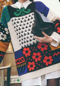 Un pull en tricot avec un design en patchwork comprenant des motifs floraux, géométriques et de feuilles en rouge, noir, bleu, vert et beige. Sac à main noir tenu.