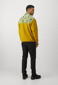 Gul fleece-jakke med et mønstret blått og hvitt bryst, lange ermer, stående krage, sammen med svarte cargo-bukser og mørke sko.