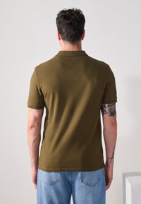 Camiseta polo verde oliva con mangas cortas, textura suave, diseño ajustado y un collar sutil. Vista trasera que muestra detalles de costura.
