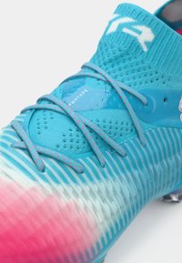 Gros plan sur une chaussure de football dégradé bleu et rose avec texture en maille, lacets et logo blanc sur le col de la cheville.