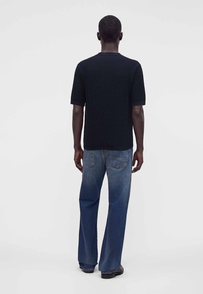 Homme debout, dos à nous, portant une chemise noire à manches courtes, un jean bleu délavé et des chaussures noires sur un fond clair uni.