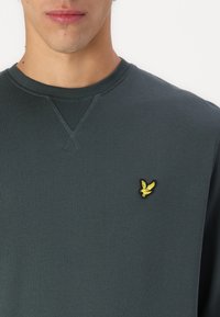 Felpa verde scuro in cotone texturizzato con colletto a girocollo e logo di un uccello ricamato giallo sul lato sinistro del petto.