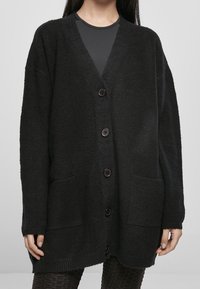 Cardigan noir tricoté avec des manches longues, décolleté en V, cinq boutons, deux poches avant et un tissu texturé.