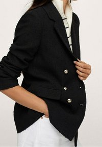 Czarna, teksturowana marynarka o podwójnym zapięciu, ze złotymi guzikami i dopasowanym krojem, noszona na białym swetrze w paski. Widoczne detale na rękawach.