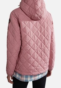 Růžový prošívaný kabát s kapucí, charakterizovaný diamantovým prošíváním a kulatým logem na levém rameni, vrstvený přes kostkovanou košili.