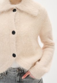 Mekan izuzetno mekan i fluffy bež kardigan s crnim dugmadima nosi se preko bijele majice, a upareno je s sivim trapericama, dok jedna ruka odmara u džepu.