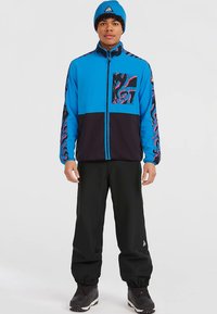 Casaco azul e preto com fecho de correr, com bolso no peito padrão; combinado com calças pretas e um gorro de malha azul. Apresenta um design geométrico nas mangas.