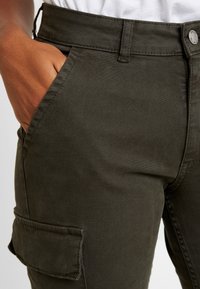 Pantalons cargo vert olive foncé en tissu texturé, avec des poches latérales et une taille ajustée avec fermeture à bouton en métal.