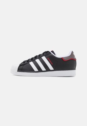 Black sneaker with white three stripes, rubber shell toe, and white sole. Features a red detail on the side marked "SUPERSTAR."